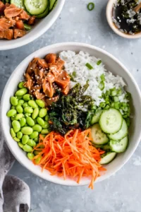 Assemble poke bowls with sushi rice, edamame, cucumber, carrot, seaweed salad, and protein