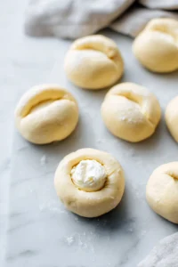 Roll the dough into small balls, then flatten each ball and place a small spoonful of the cream cheese filling in the center. Fold the dough over the filling and seal it well.