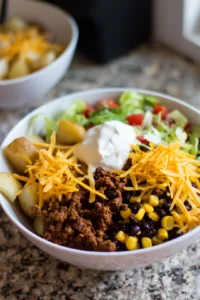 Assemble the taco bowls by layering potatoes, seasoned ground beef, black beans, corn, shredded lettuce, and cheddar cheese. Top with a dollop of sour cream and serve immediately.