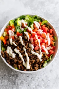 Assemble your burger bowls with lettuce, cooked ground beef, diced tomatoes, and a drizzle of sauce on top.